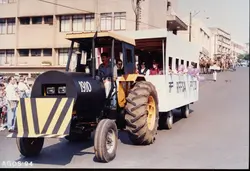 Uma fotografia colorida que mostra um veículo adaptado, possivelmente um trator decorado como um trem, participando de um desfile ou evento público. O veículo exibe a inscrição 'RFFSA' e o número '1910', sugerindo uma homenagem à Rede Ferroviária Federal, com pessoas visíveis na estrutura e ao fundo em uma área urbana.