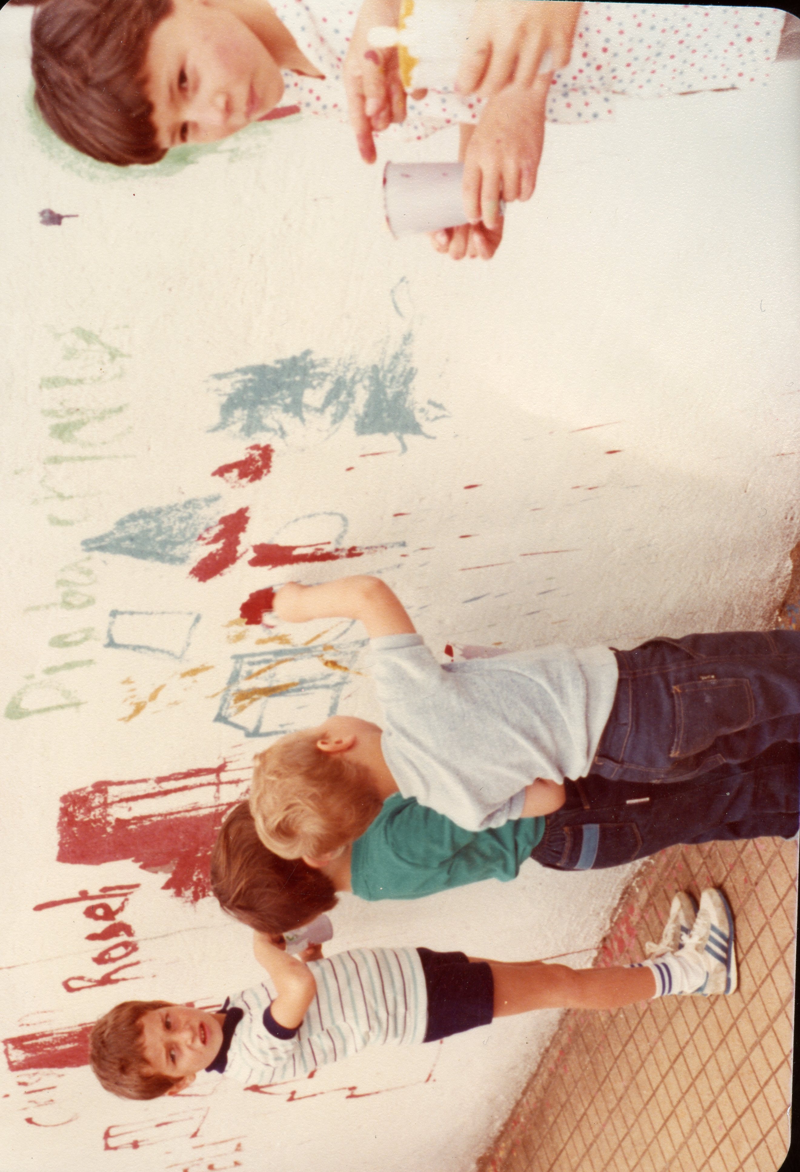 Uma fotografia colorida que captura um grupo de crianças pintando uma parede externa, possivelmente em um ambiente escolar ou recreativo. A cena reflete uma atividade lúdica típica das décadas de 1980 ou 1990, com as crianças usando roupas casuais da época.