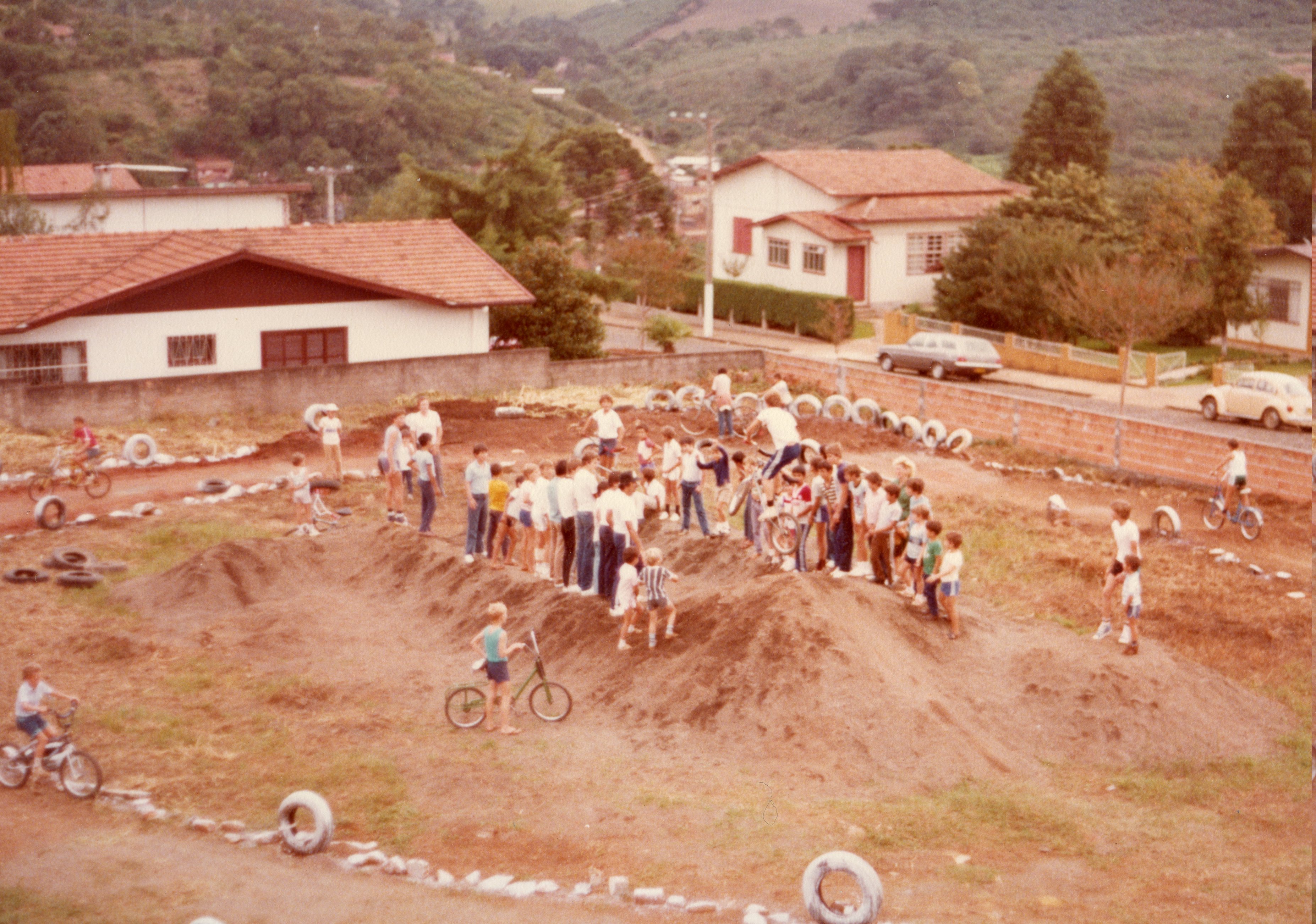Uma cena ao ar livre mostrando um grupo de crianças e jovens reunidos em um terreno baldio adaptado como pista de ciclismo improvisada, com montes de terra e pneus como obstáculos. A imagem reflete o cotidiano recreativo de uma pequena cidade brasileira, possivelmente na década de 1980, com casas de arquitetura típica da região sul ao fundo.