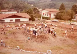 Uma cena ao ar livre mostrando um grupo de crianças e jovens reunidos em um terreno baldio adaptado como pista de ciclismo improvisada, com montes de terra e pneus como obstáculos. A imagem reflete o cotidiano recreativo de uma pequena cidade brasileira, possivelmente na década de 1980, com casas de arquitetura típica da região sul ao fundo.