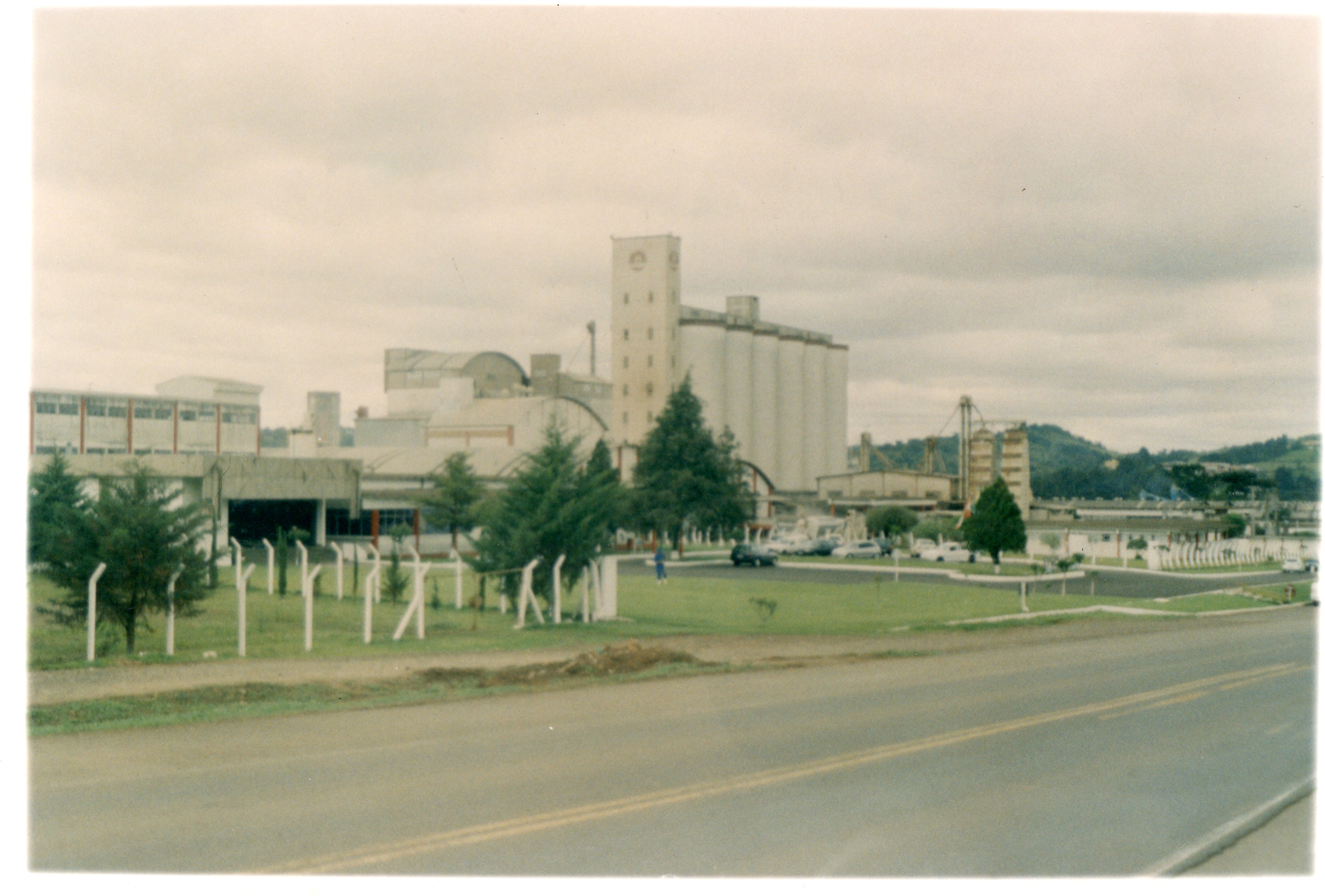 Uma fotografia colorida de um complexo industrial, possivelmente uma unidade de processamento de grãos ou agroindústria, com grandes silos e estruturas fabris. A cena apresenta uma área externa com gramado, árvores e uma estrada pavimentada em primeiro plano, sugerindo um ambiente rural ou periurbano típico de cidades do interior brasileiro.
