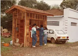 Uma fotografia colorida que mostra um grupo de jovens interagindo em um quiosque de madeira recém-construído. Ao lado, um carro Fiat 147 branco estacionado sugere uma cena típica do cotidiano brasileiro entre o final da década de 1980 e início da de 1990.