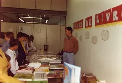 Uma cena interna capturando uma Feira do Livro escolar ou comunitária, com um grupo de jovens observando livros expostos em mesas. O ambiente apresenta decoração temática nas paredes e um homem adulto supervisionando a atividade, datado de maio de 1995.