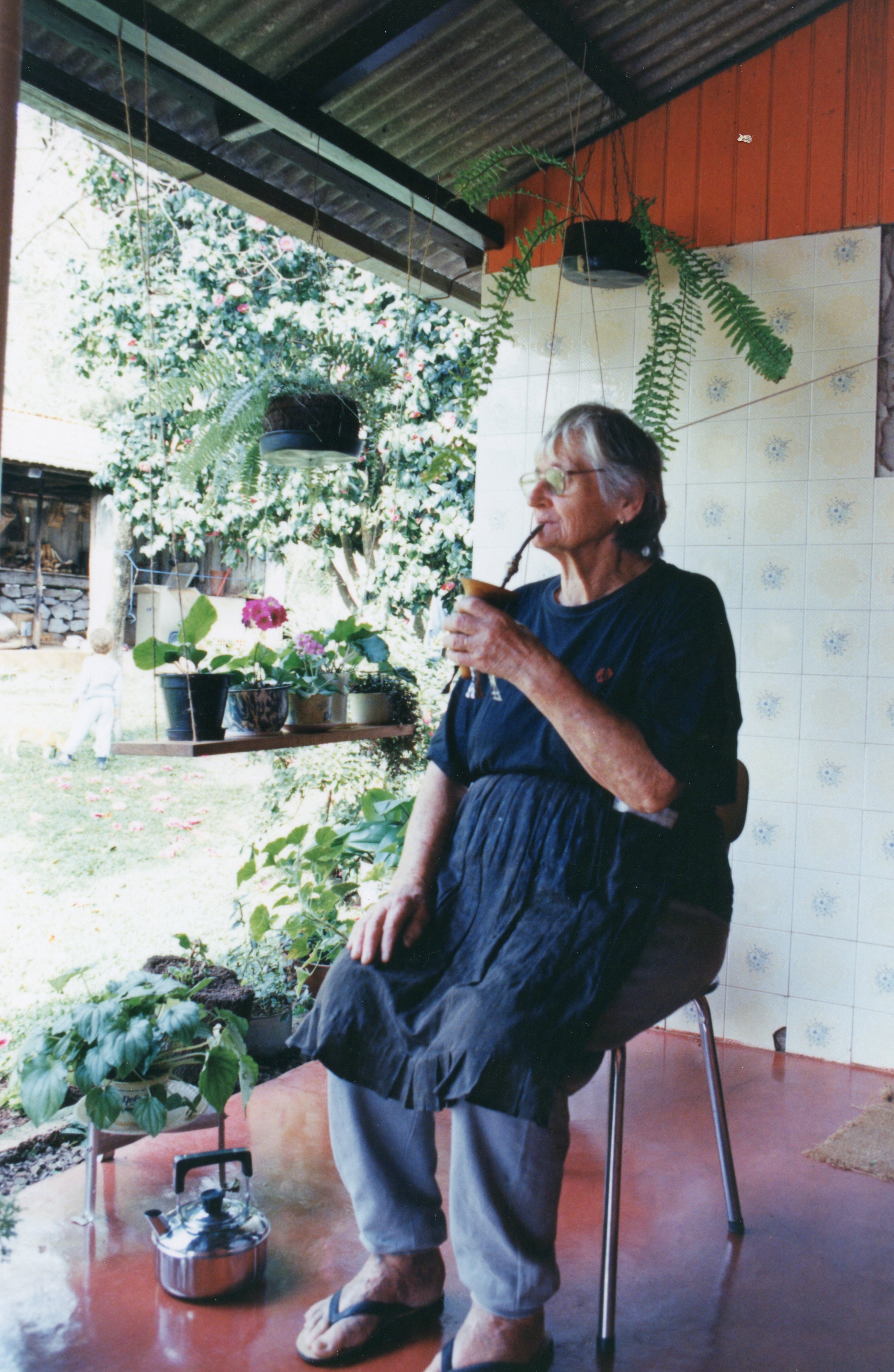 Uma senhora idosa está sentada em uma varanda, tomando chimarrão, vestindo roupas simples e um avental. O ambiente é uma residência rural ou suburbana com plantas pendentes e uma criança pequena ao fundo, sugerindo um momento cotidiano de lazer em uma região de colonização sulista.