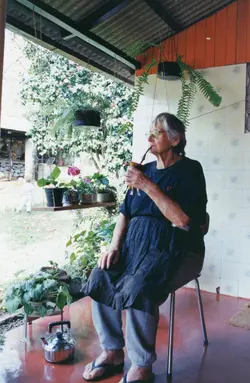 Uma senhora idosa está sentada em uma varanda, tomando chimarrão, vestindo roupas simples e um avental. O ambiente é uma residência rural ou suburbana com plantas pendentes e uma criança pequena ao fundo, sugerindo um momento cotidiano de lazer em uma região de colonização sulista.