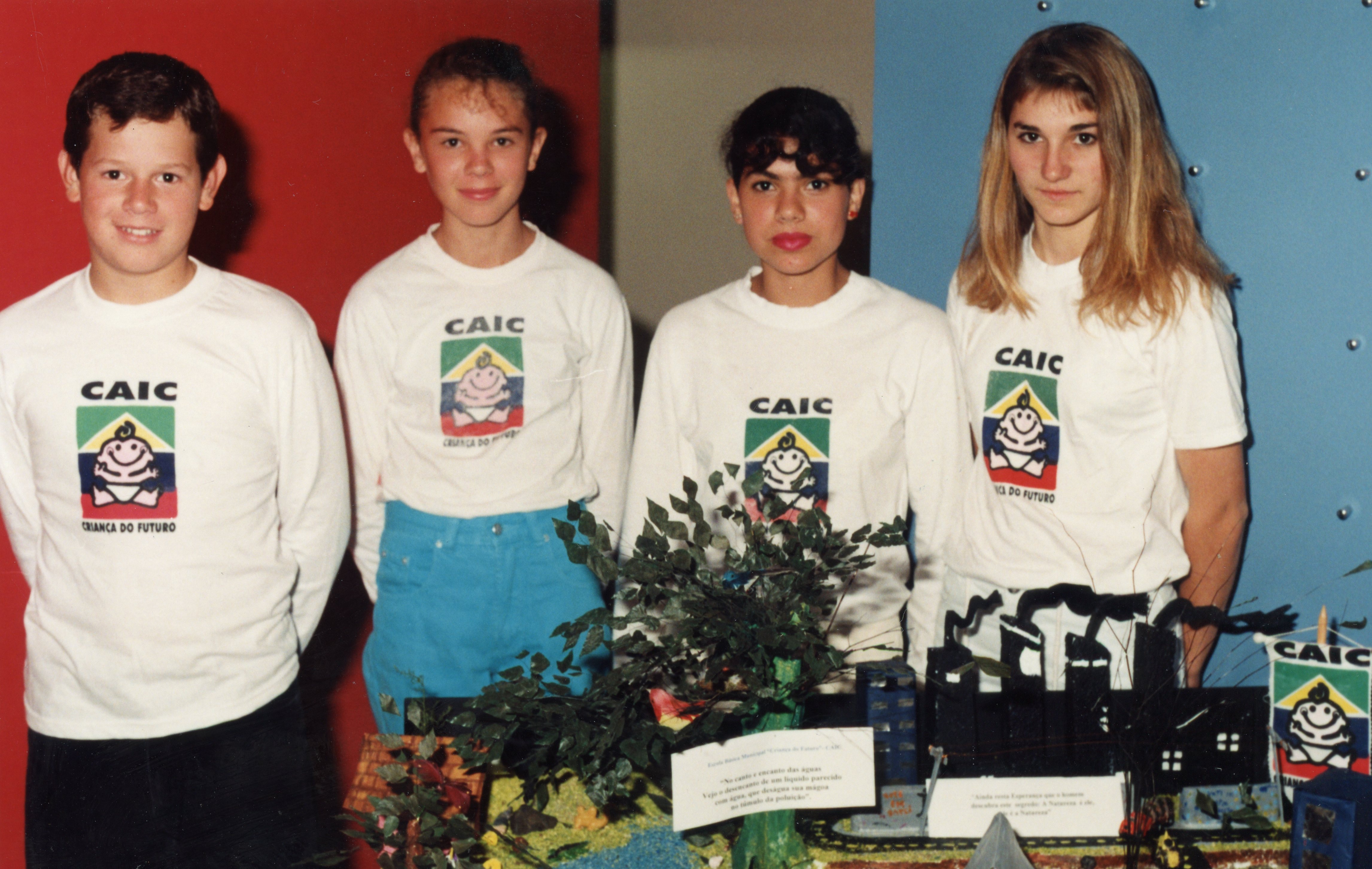 Uma fotografia de quatro jovens estudantes vestindo camisetas brancas com o logotipo do projeto CAIC (Criança do Futuro), provavelmente em uma feira escolar ou evento educacional. Eles estão posicionados atrás de uma maquete que aborda temas ambientais, sugerindo uma atividade pedagógica focada em conscientização ecológica, típica da década de 1990 no Brasil.