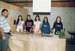 Uma feira de ciências escolar onde um professor e cinco alunas apresentam um projeto sobre desenvolvimento fetal. O ambiente é simples, com paredes de tijolos aparentes e cartazes informativos, sugerindo uma escola pública brasileira na década de 1990.
