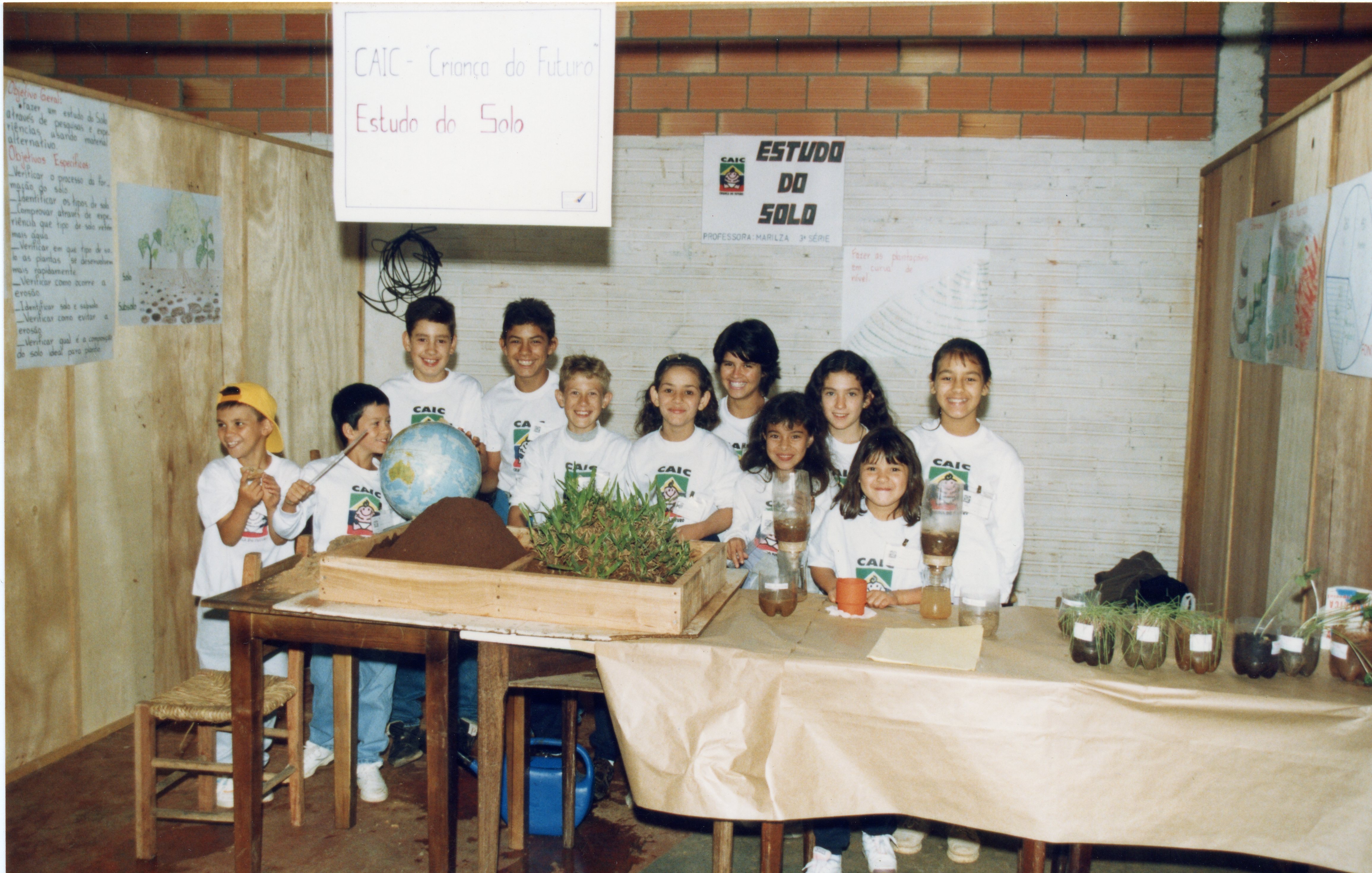 Uma fotografia escolar mostrando um grupo de crianças em uma feira de ciências com o tema 'Estudo do Solo'. Os alunos vestem camisetas padronizadas do CAIC e estão atrás de uma mesa com experimentos práticos, incluindo um globo terrestre e amostras de plantas em garrafas PET, sugerindo uma atividade pedagógica típica dos anos 90.