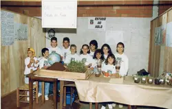 Uma fotografia escolar mostrando um grupo de crianças em uma feira de ciências com o tema 'Estudo do Solo'. Os alunos vestem camisetas padronizadas do CAIC e estão atrás de uma mesa com experimentos práticos, incluindo um globo terrestre e amostras de plantas em garrafas PET, sugerindo uma atividade pedagógica típica dos anos 90.