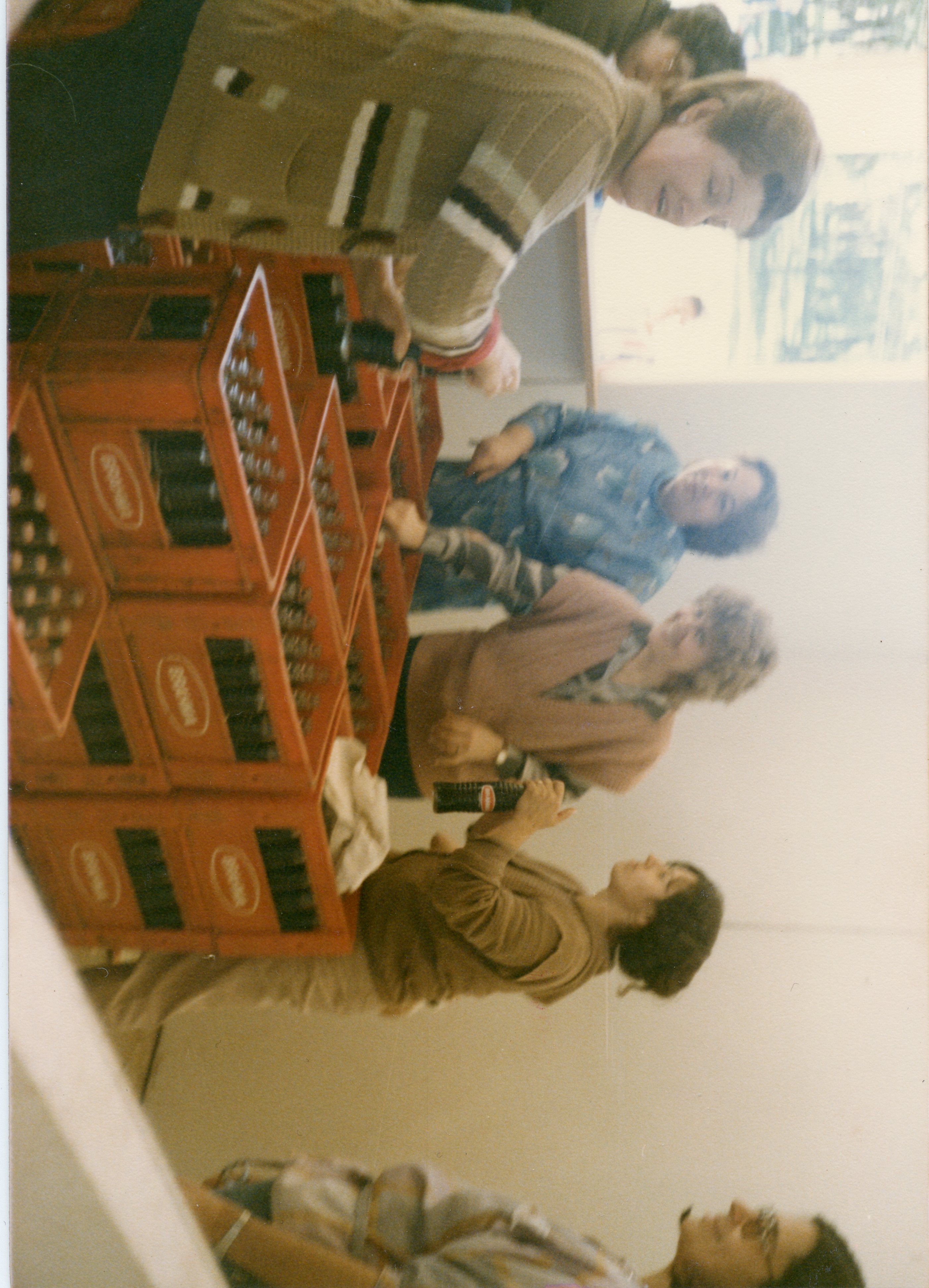 Uma fotografia que captura um grupo de mulheres interagindo em um ambiente interno, possivelmente um comércio ou depósito de bebidas. Elas estão organizando ou manuseando engradados de cerveja da marca Brahma, sugerindo um contexto de trabalho ou preparação para um evento social, com vestimentas típicas das décadas de 1980 ou 1990.