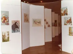 Uma exposição de arte organizada em um espaço interno com divisórias brancas dispostas em zigue-zague. Diversas pinturas, incluindo naturezas-mortas, retratos de animais e paisagens, estão penduradas nas paredes, sugerindo um evento cultural ou galeria de arte de meados da década de 1980.