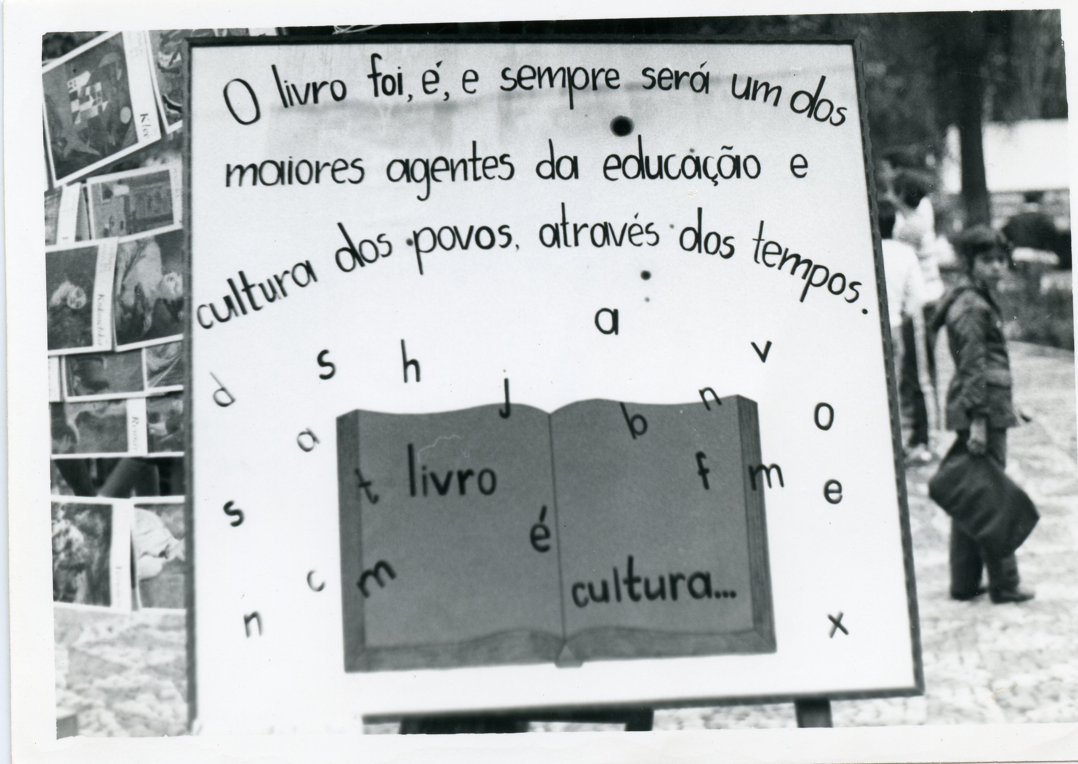 Uma fotografia em preto e branco exibindo um cartaz educativo sobre a importância dos livros, provavelmente parte de uma feira cultural ou evento escolar. O cartaz contém uma mensagem escrita à mão e ilustrações de letras espalhadas, com uma criança visível ao fundo em um ambiente externo.
