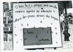 Uma fotografia em preto e branco exibindo um cartaz educativo sobre a importância dos livros, provavelmente parte de uma feira cultural ou evento escolar. O cartaz contém uma mensagem escrita à mão e ilustrações de letras espalhadas, com uma criança visível ao fundo em um ambiente externo.