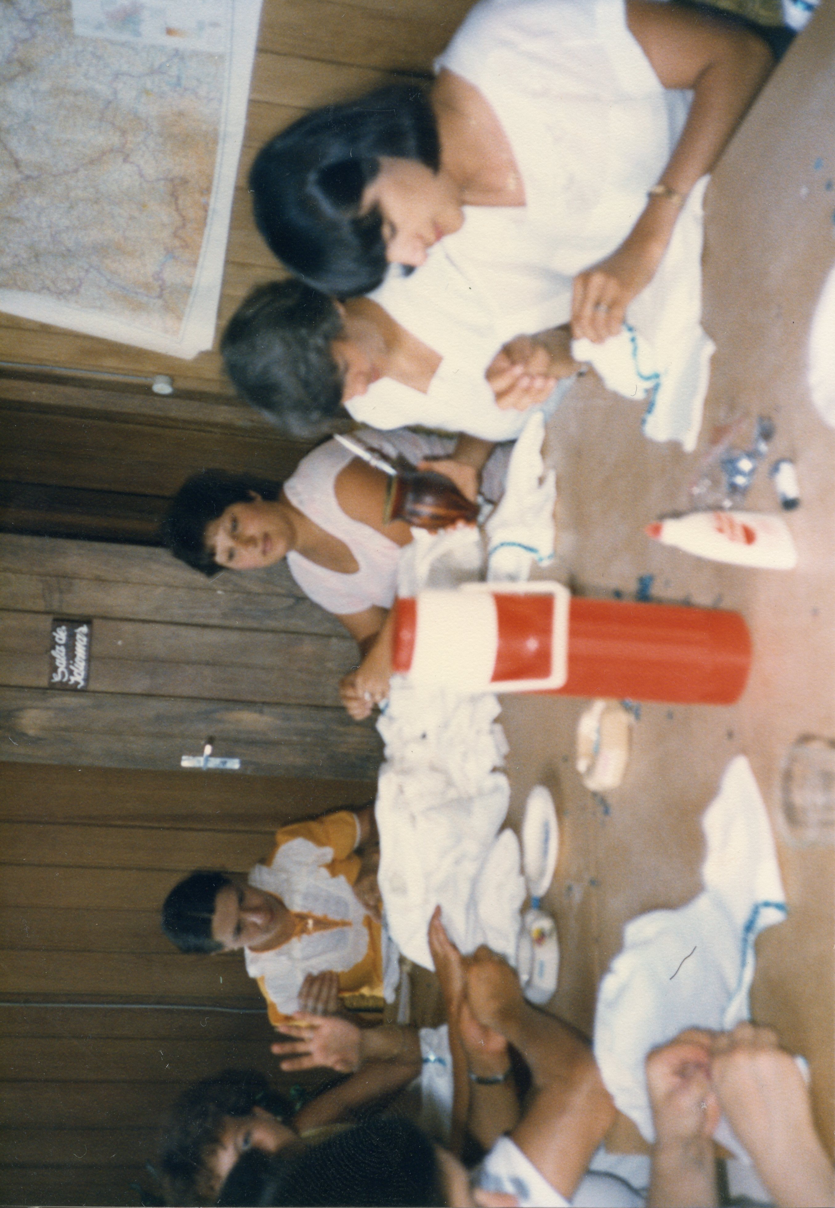 Uma fotografia colorida de um grupo de mulheres reunidas em torno de uma mesa de madeira, possivelmente em um ambiente escolar ou comunitário, como sugere a placa 'Sala de Idiomas'. Elas parecem estar envolvidas em uma atividade manual ou de artesanato, acompanhadas por uma garrafa térmica de chimarrão, refletindo um costume social comum no sul do Brasil.