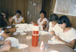 Uma fotografia colorida de um grupo de mulheres reunidas em torno de uma mesa de madeira, possivelmente em um ambiente escolar ou comunitário, como sugere a placa 'Sala de Idiomas'. Elas parecem estar envolvidas em uma atividade manual ou de artesanato, acompanhadas por uma garrafa térmica de chimarrão, refletindo um costume social comum no sul do Brasil.