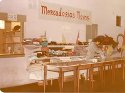 Uma fotografia interna que mostra um espaço de vendas improvisado, possivelmente um bazar ou feira comunitária, com cartazes escritos à mão indicando 'Mercadorias Novas'. Uma pessoa organiza roupas sobre mesas simples, em um ambiente que parece ser um salão paroquial ou comunitário, sugerindo uma atividade de arrecadação ou comércio local de meados da década de 1980.
