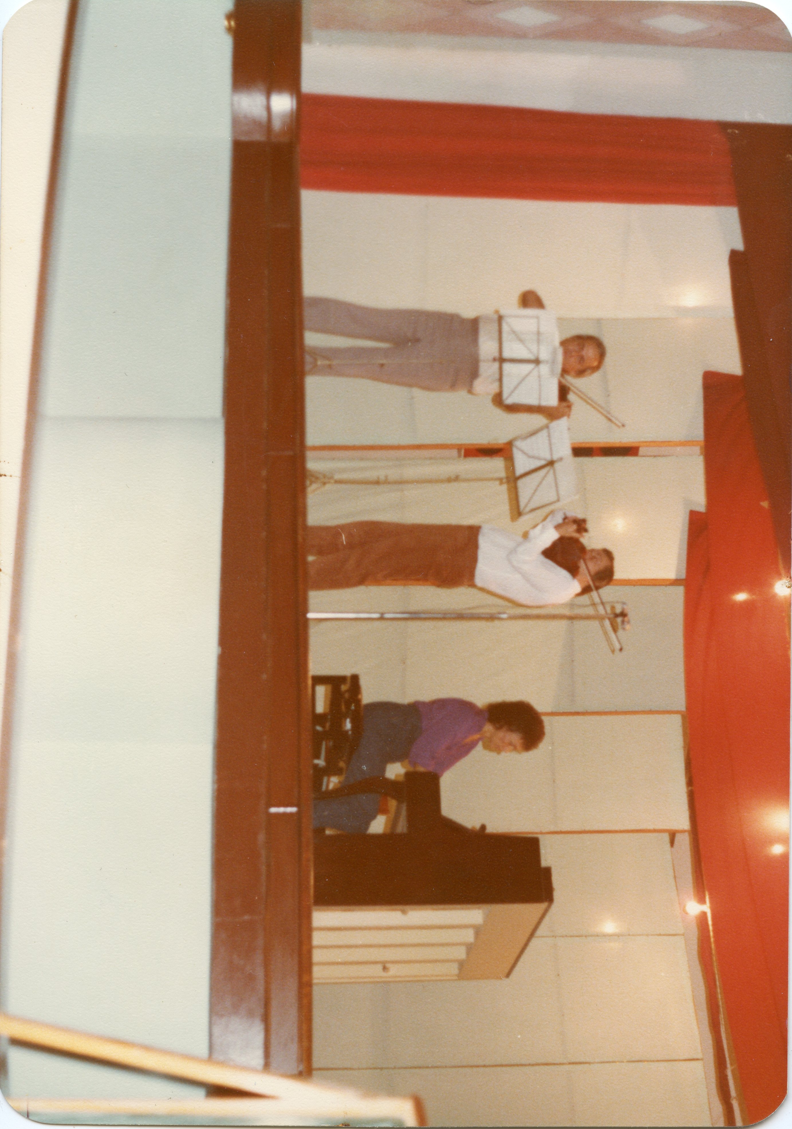 Uma fotografia capturada de um ângulo lateral mostrando três músicos em um palco, possivelmente durante um ensaio ou apresentação escolar. O grupo inclui dois violinistas e uma pessoa tocando piano, com cortinas vermelhas ao fundo, sugerindo um ambiente de auditório típico de meados da década de 1980.