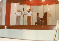 Uma fotografia capturada de um ângulo lateral mostrando três músicos em um palco, possivelmente durante um ensaio ou apresentação escolar. O grupo inclui dois violinistas e uma pessoa tocando piano, com cortinas vermelhas ao fundo, sugerindo um ambiente de auditório típico de meados da década de 1980.