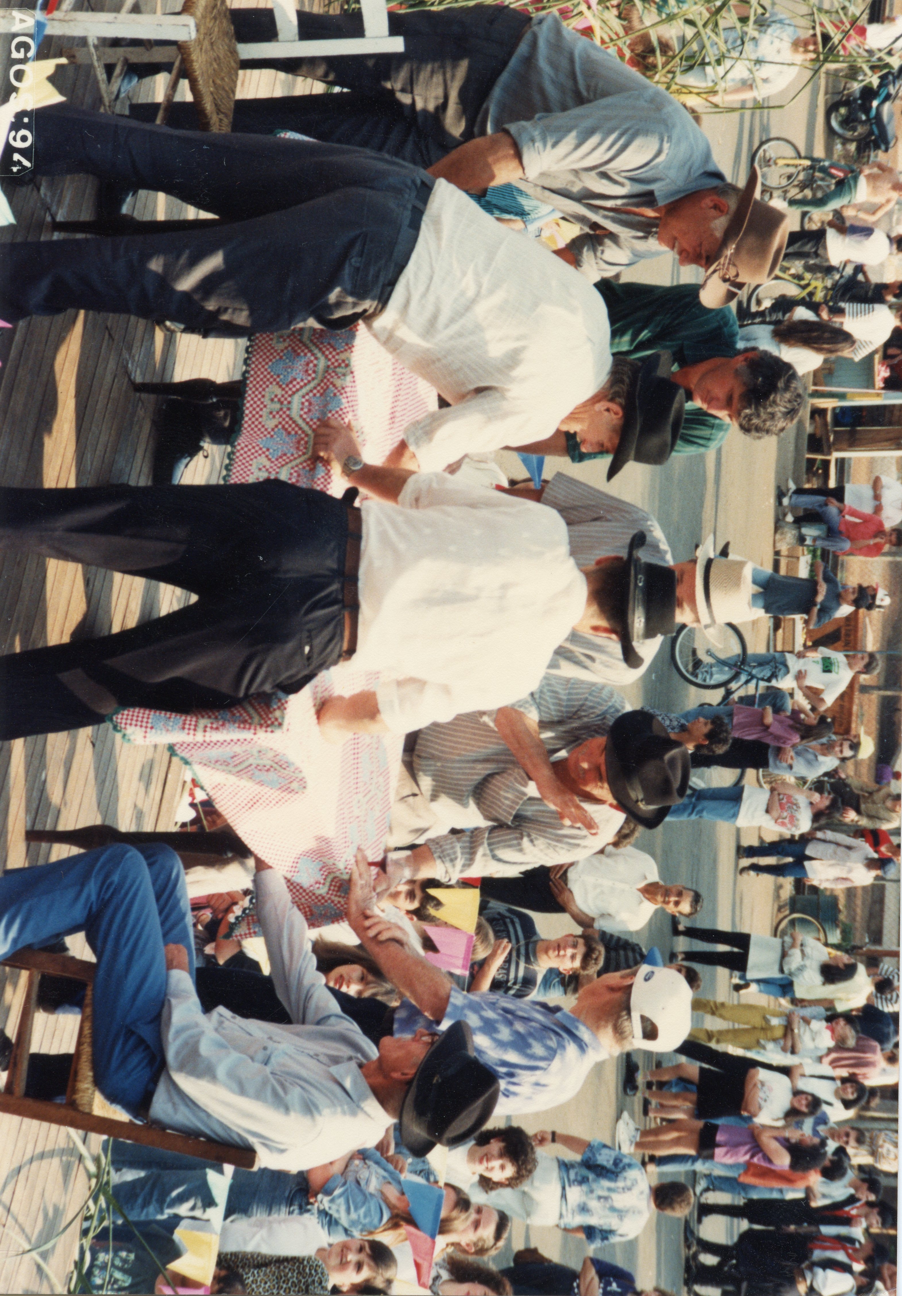 Uma cena ao ar livre mostrando um grupo de pessoas reunidas em torno de uma mesa coberta com uma toalha xadrez, possivelmente em um evento comunitário ou feira. A fotografia, datada de agosto de 1994, captura homens usando chapéus e roupas casuais, sugerindo um contexto rural ou de interior brasileiro.