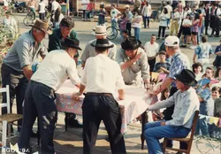 Uma cena ao ar livre mostrando um grupo de pessoas reunidas em torno de uma mesa coberta com uma toalha xadrez, possivelmente em um evento comunitário ou feira. A fotografia, datada de agosto de 1994, captura homens usando chapéus e roupas casuais, sugerindo um contexto rural ou de interior brasileiro.