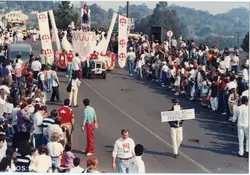 Uma fotografia de um desfile comemorativo, possivelmente em Videira, celebrando os 60 anos da empresa Perdigão. A imagem mostra uma multidão acompanhando um carro alegórico em uma via pública, com pessoas vestindo roupas típicas da década de 1990.