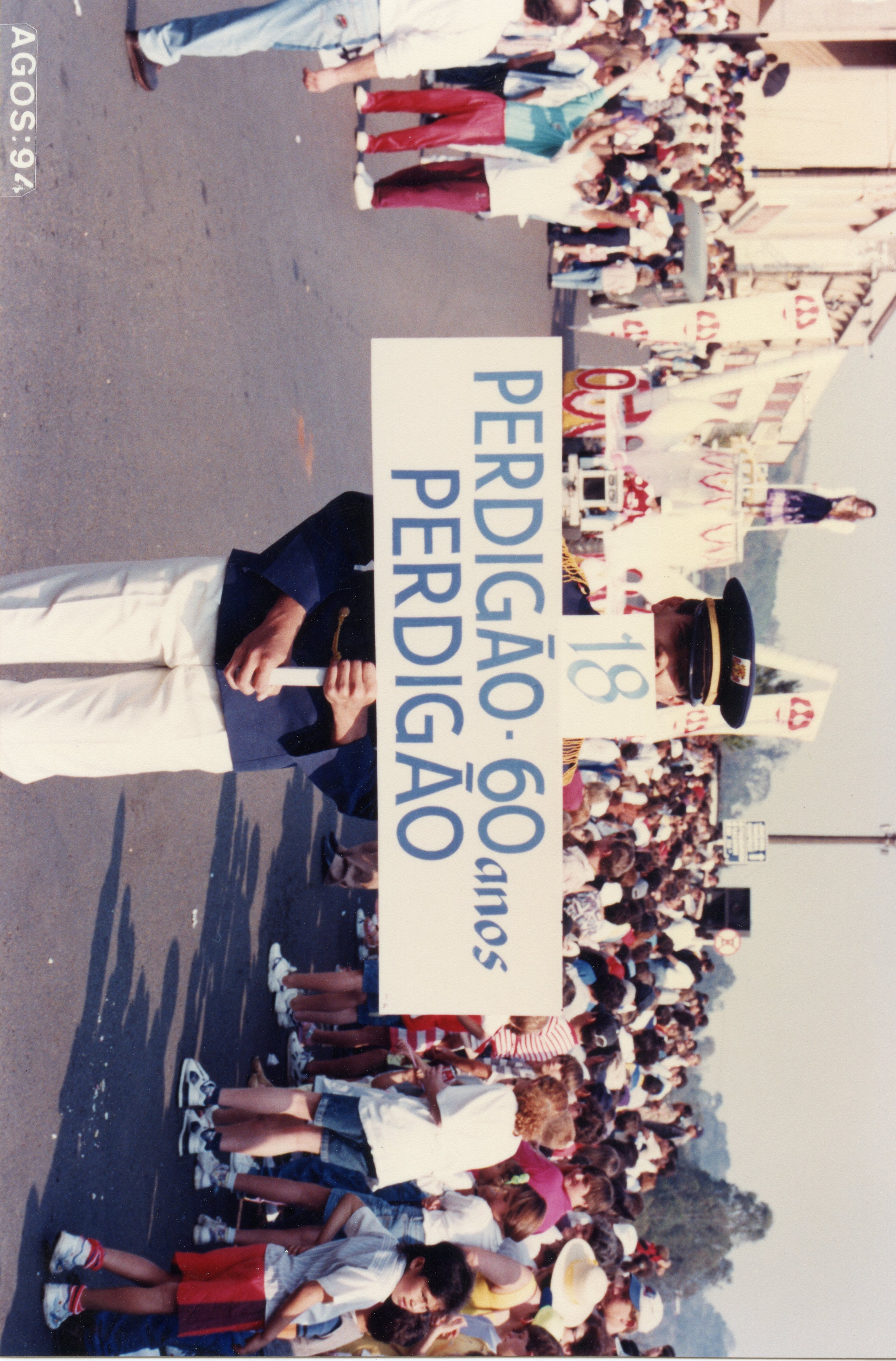 Fotografia de um desfile comemorativo em via pública, provavelmente em Videira, celebrando os 60 anos da empresa Perdigão. A imagem mostra uma multidão acompanhando o evento, com destaque para uma pessoa uniformizada segurando uma placa comemorativa datada de agosto de 1994.