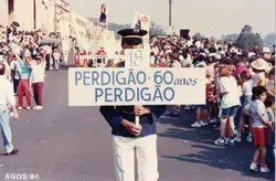 Fotografia de um desfile comemorativo em via pública, provavelmente em Videira, celebrando os 60 anos da empresa Perdigão. A imagem mostra uma multidão acompanhando o evento, com destaque para uma pessoa uniformizada segurando uma placa comemorativa datada de agosto de 1994.