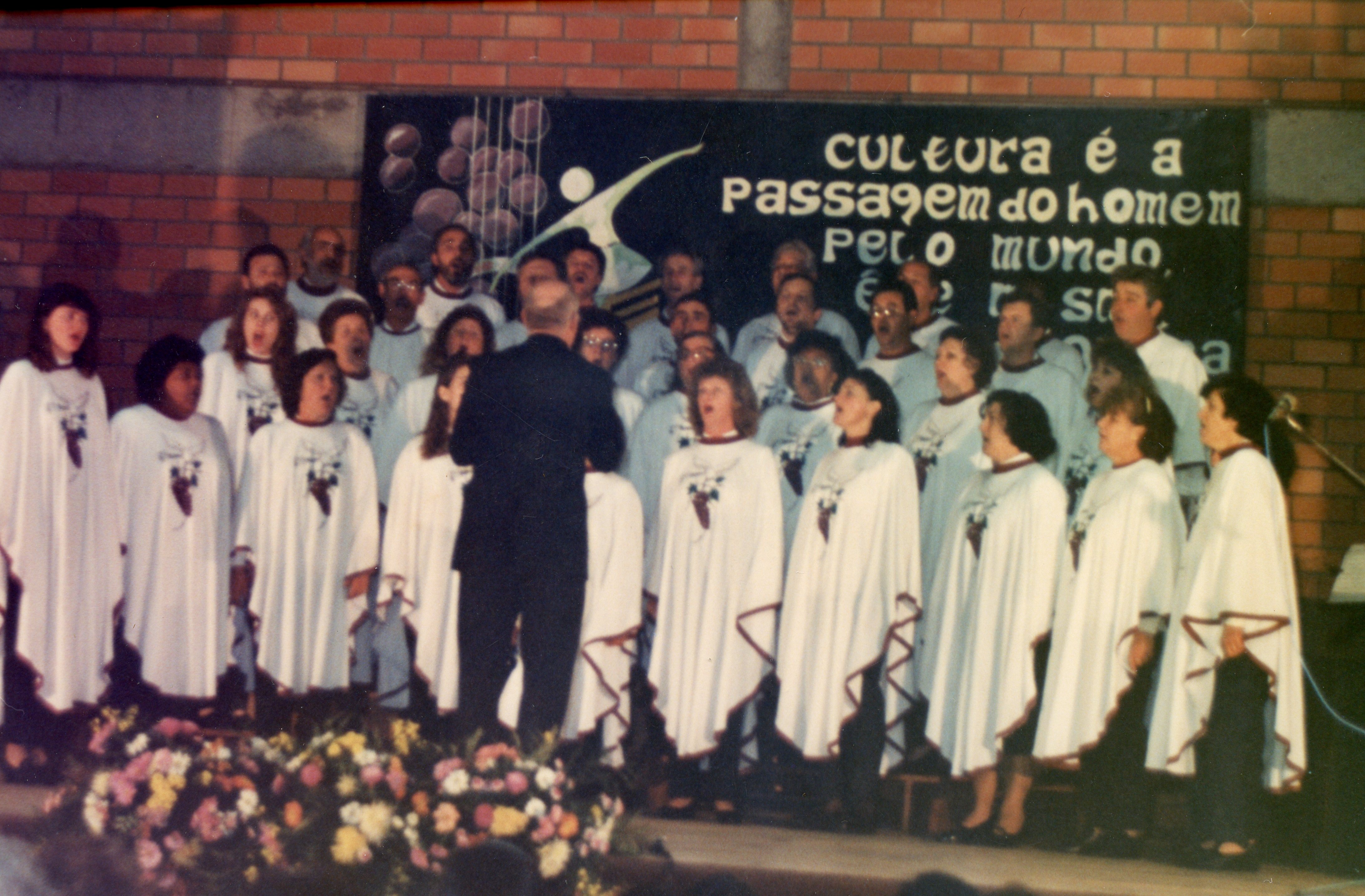 Uma fotografia de um coral apresentando-se em um ambiente interno com paredes de tijolos aparentes. O grupo, composto por homens e mulheres vestindo capas brancas com detalhes, é regido por um maestro de costas, diante de um cartaz com a frase 'Cultura é a passagem do homem pelo mundo'.