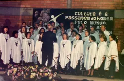 Uma fotografia de um coral apresentando-se em um ambiente interno com paredes de tijolos aparentes. O grupo, composto por homens e mulheres vestindo capas brancas com detalhes, é regido por um maestro de costas, diante de um cartaz com a frase 'Cultura é a passagem do homem pelo mundo'.