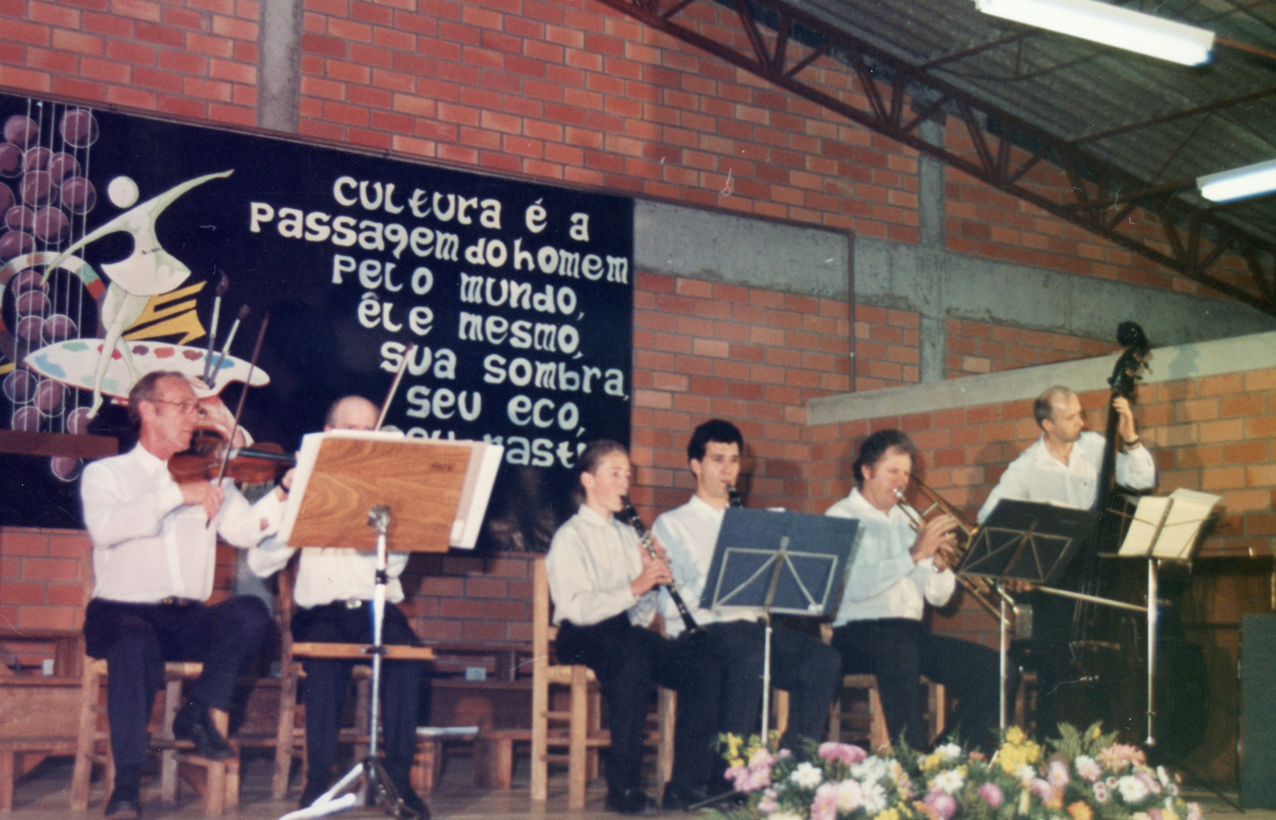 Uma pequena orquestra ou conjunto musical apresenta-se em um ambiente interno com paredes de tijolos aparentes. Ao fundo, um cartaz com uma frase sobre cultura sugere um evento cultural ou escolar, possivelmente em uma comunidade brasileira.