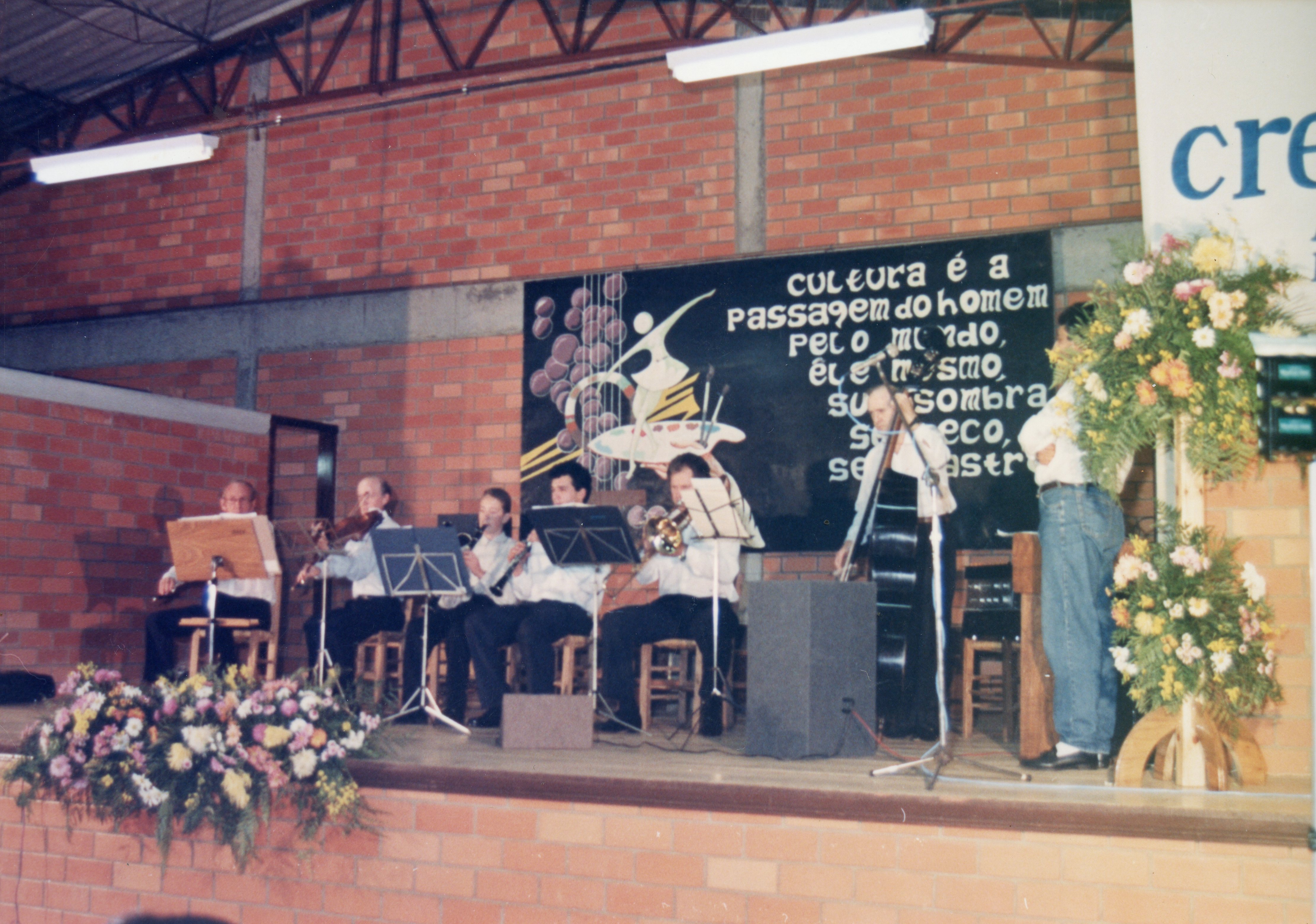 Uma pequena orquestra ou conjunto musical apresenta-se em um palco com parede de tijolos aparentes, decorado com um painel artístico contendo uma frase sobre cultura. A cena sugere um evento cultural ou escolar, possivelmente nas décadas de 1980 ou 1990, dado o estilo das vestimentas e a qualidade da fotografia.