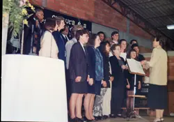 Uma fotografia de um coral apresentando-se em um ambiente interno com paredes de tijolos aparentes. O grupo, composto por homens e mulheres vestidos formalmente, é regido por uma mulher de costas para a câmera, sugerindo um evento cultural ou religioso típico de comunidades brasileiras nas décadas de 1980 ou 1990.