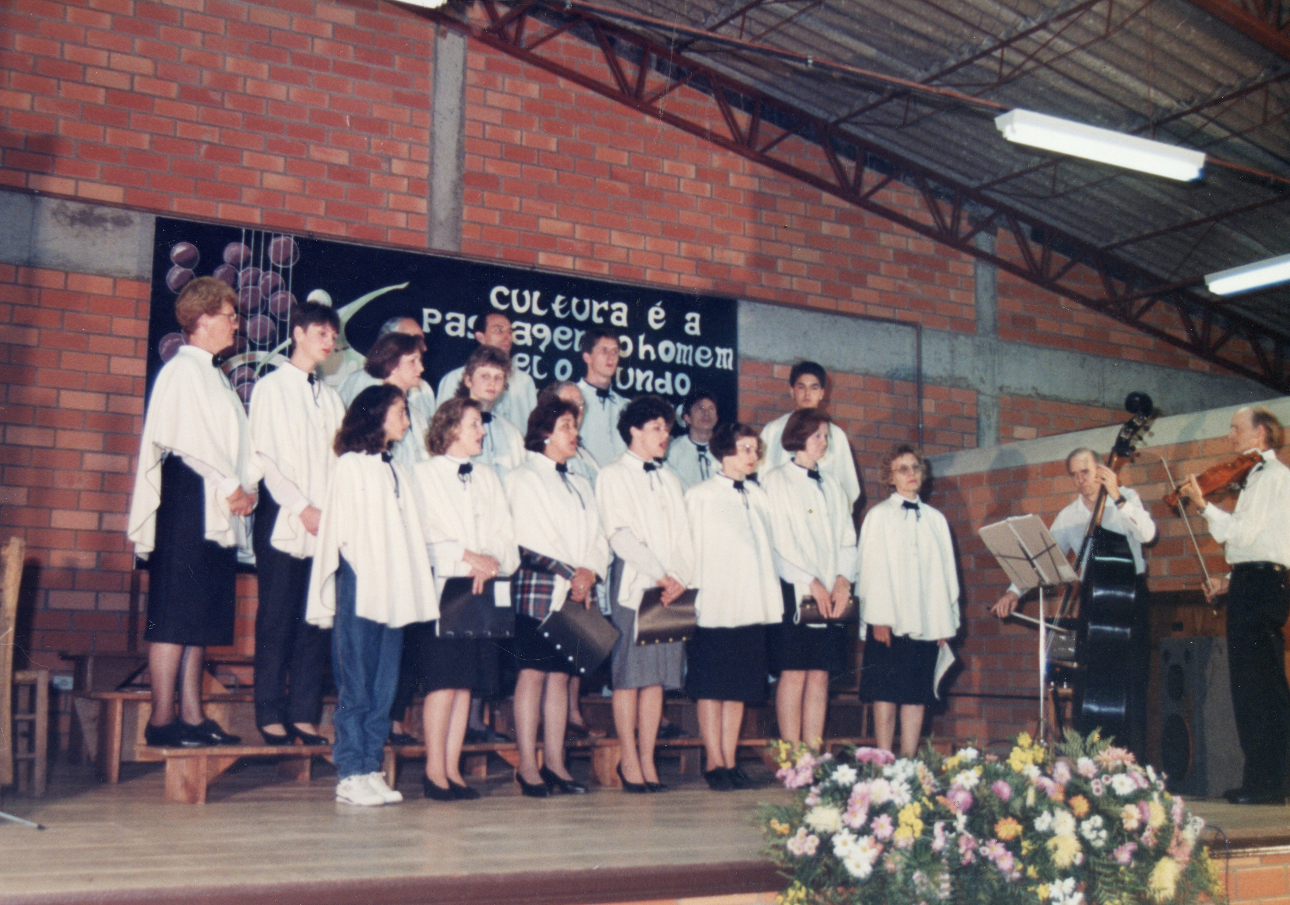 Uma fotografia de um coral apresentando-se em um palco com fundo de tijolos aparentes, possivelmente em um evento cultural ou escolar. Os integrantes vestem capas brancas e roupas escuras, acompanhados por músicos com violino e contrabaixo, sugerindo uma cena típica de celebrações comunitárias ou escolares das décadas de 1980 ou 1990.