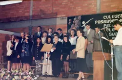 Uma fotografia de um grupo de pessoas em um evento formal, possivelmente um coral ou cerimônia cultural, em um ambiente interno com paredes de tijolos aparentes. O contexto sugere um evento comunitário ou escolar, com um cartaz ao fundo mencionando 'cultura' e 'passagem', indicando um registro de atividade social ou educacional, provavelmente da década de 1990.