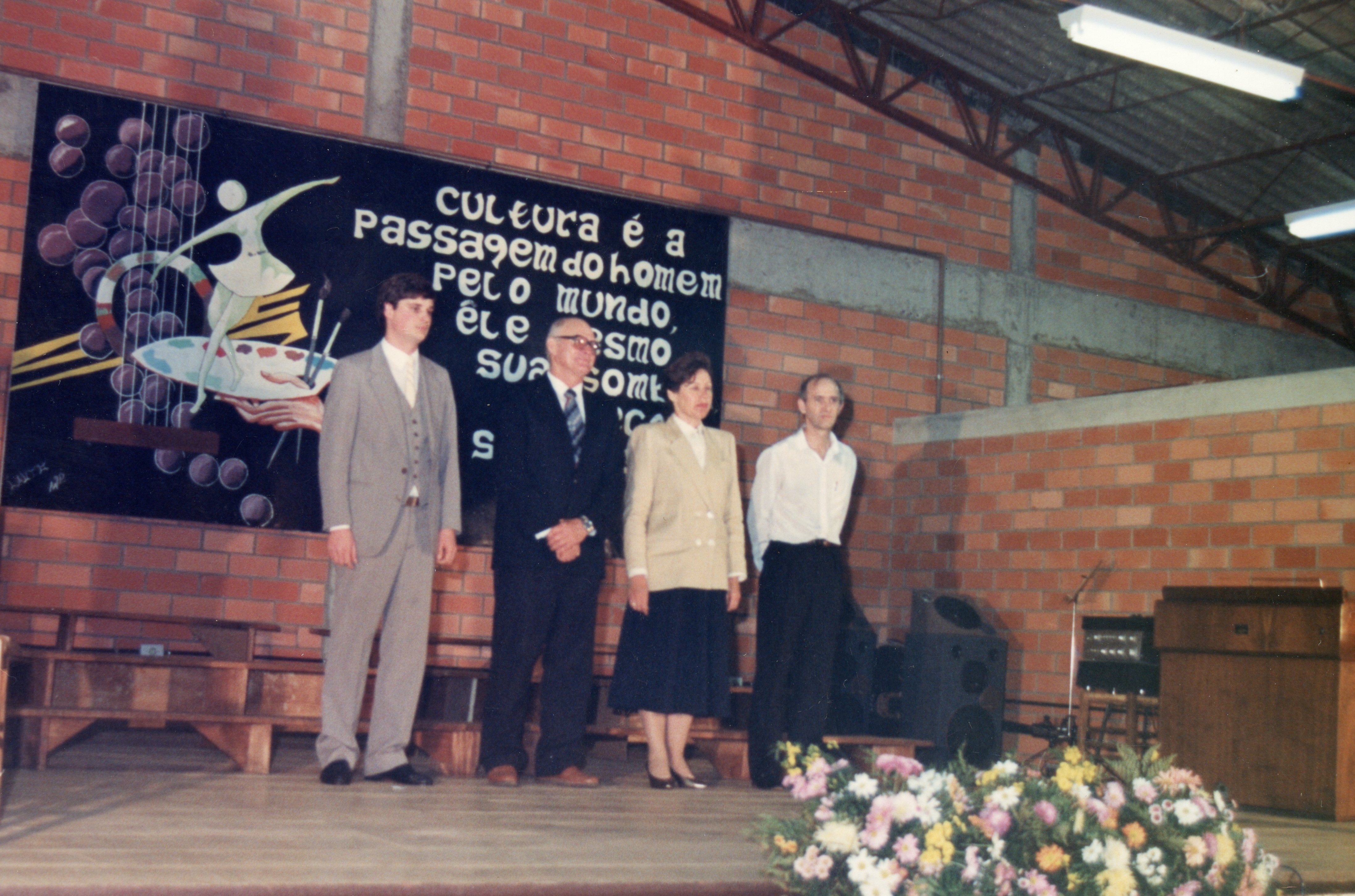 Uma fotografia de um evento formal realizado em um ambiente com paredes de tijolos aparentes, possivelmente um centro comunitário ou escola. Quatro pessoas estão em pé no palco, diante de um painel artístico com uma frase sobre cultura, sugerindo uma cerimônia de formatura ou evento cultural ocorrido provavelmente nas décadas de 1980 ou 1990.