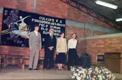 Uma fotografia de um evento formal realizado em um ambiente com paredes de tijolos aparentes, possivelmente um centro comunitário ou escola. Quatro pessoas estão em pé no palco, diante de um painel artístico com uma frase sobre cultura, sugerindo uma cerimônia de formatura ou evento cultural ocorrido provavelmente nas décadas de 1980 ou 1990.