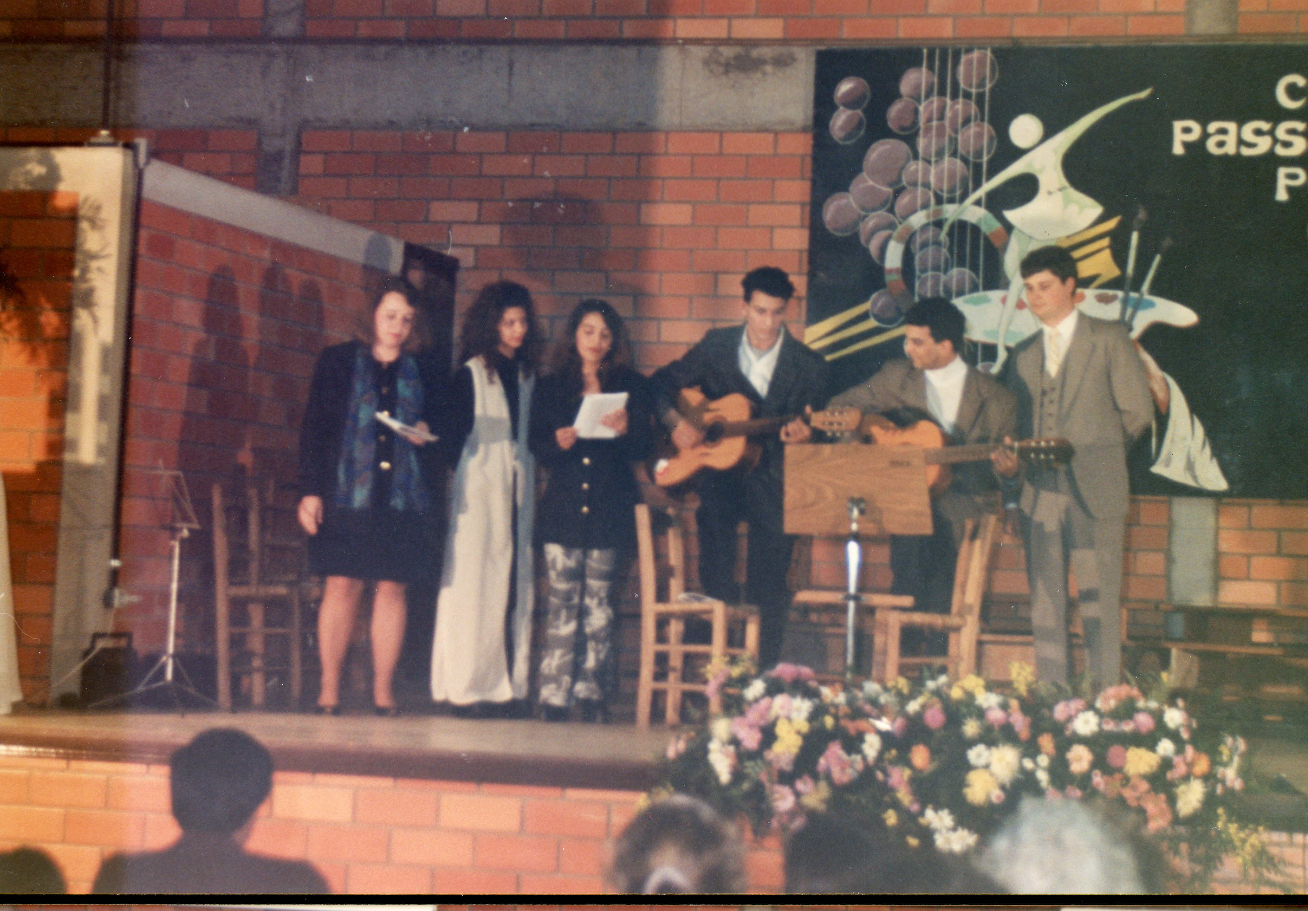 Uma fotografia de um evento cultural ou escolar em um palco com parede de tijolos aparentes, apresentando um grupo de pessoas cantando e tocando violão. O estilo das vestimentas e a qualidade da imagem sugerem um evento ocorrido provavelmente na década de 1990 em um contexto comunitário ou escolar.