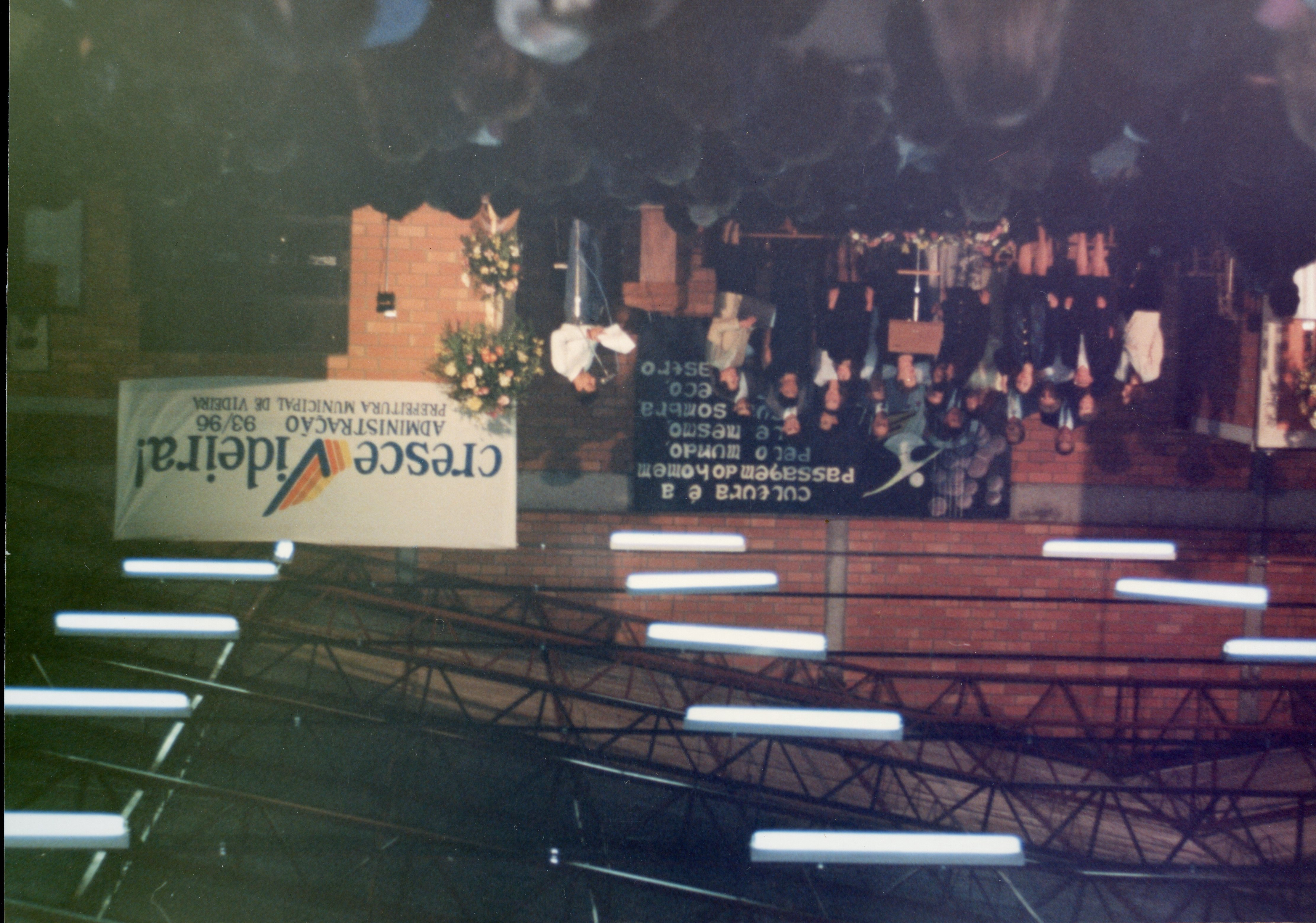 Uma fotografia de um evento público em um ambiente interno, provavelmente um ginásio ou centro cultural em Videira, Santa Catarina. O banner indica a administração municipal de 1993-1996, sugerindo um evento oficial ou cultural daquela época.
