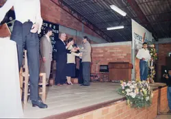 Uma cerimônia de entrega de prêmios ou homenagens ocorrendo em um palco com estrutura de tijolos aparentes e teto metálico. O evento parece ser oficial ou comunitário, com a presença de um banner com os dizeres 'Cresce Videira', sugerindo uma atividade administrativa ou cívica na cidade de Videira, Santa Catarina, provavelmente na década de 1990.