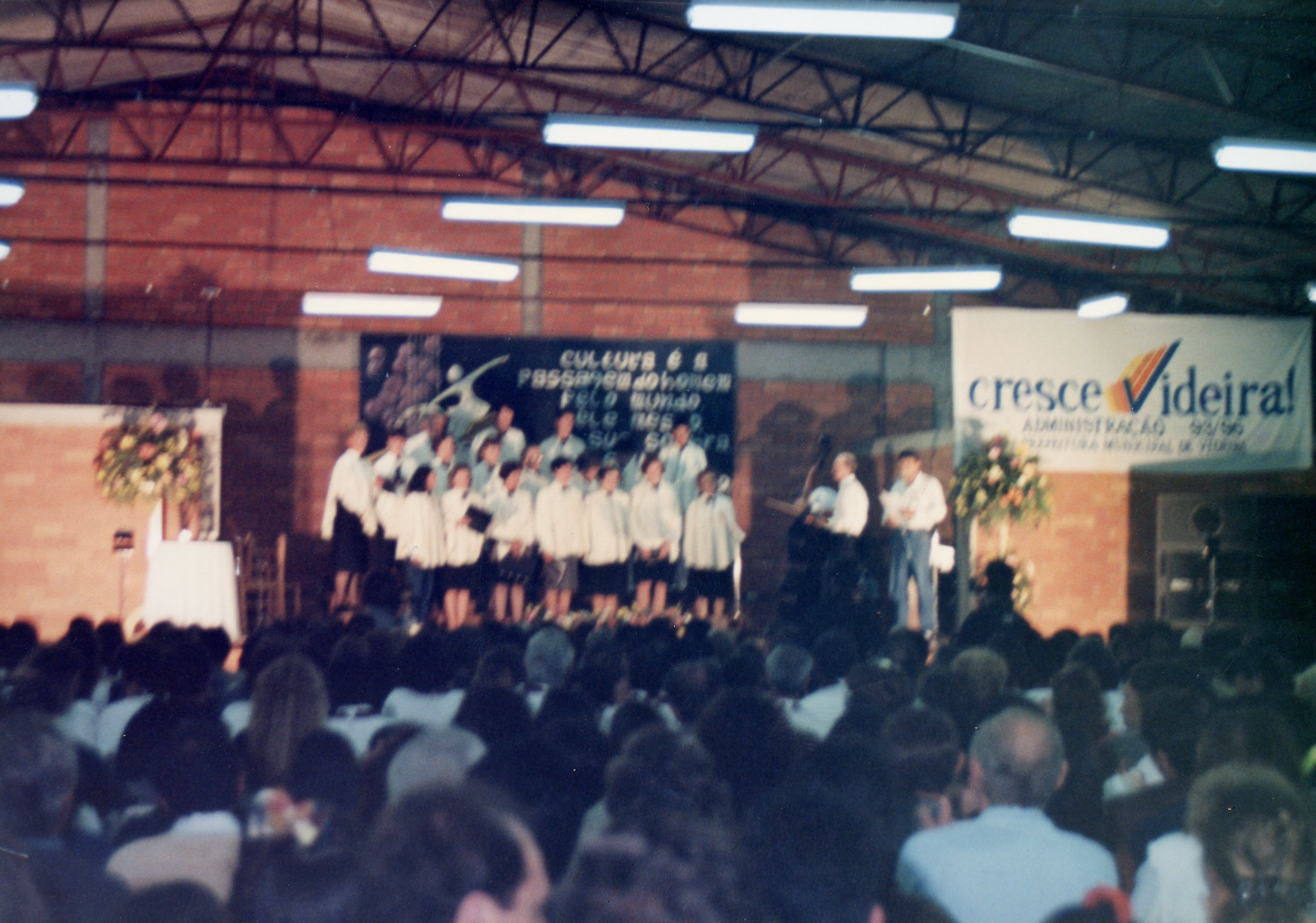 Uma fotografia de um evento público em um ginásio ou salão coberto, com um coral ou grupo musical se apresentando no palco. O banner ao fundo menciona 'Cresce Videira! Administração 93/96', indicando um evento oficial na cidade de Videira, Santa Catarina, durante meados da década de 1990.