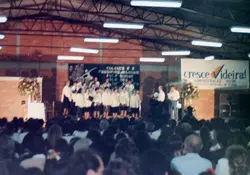 Uma fotografia de um evento público em um ginásio ou salão coberto, com um coral ou grupo musical se apresentando no palco. O banner ao fundo menciona 'Cresce Videira! Administração 93/96', indicando um evento oficial na cidade de Videira, Santa Catarina, durante meados da década de 1990.