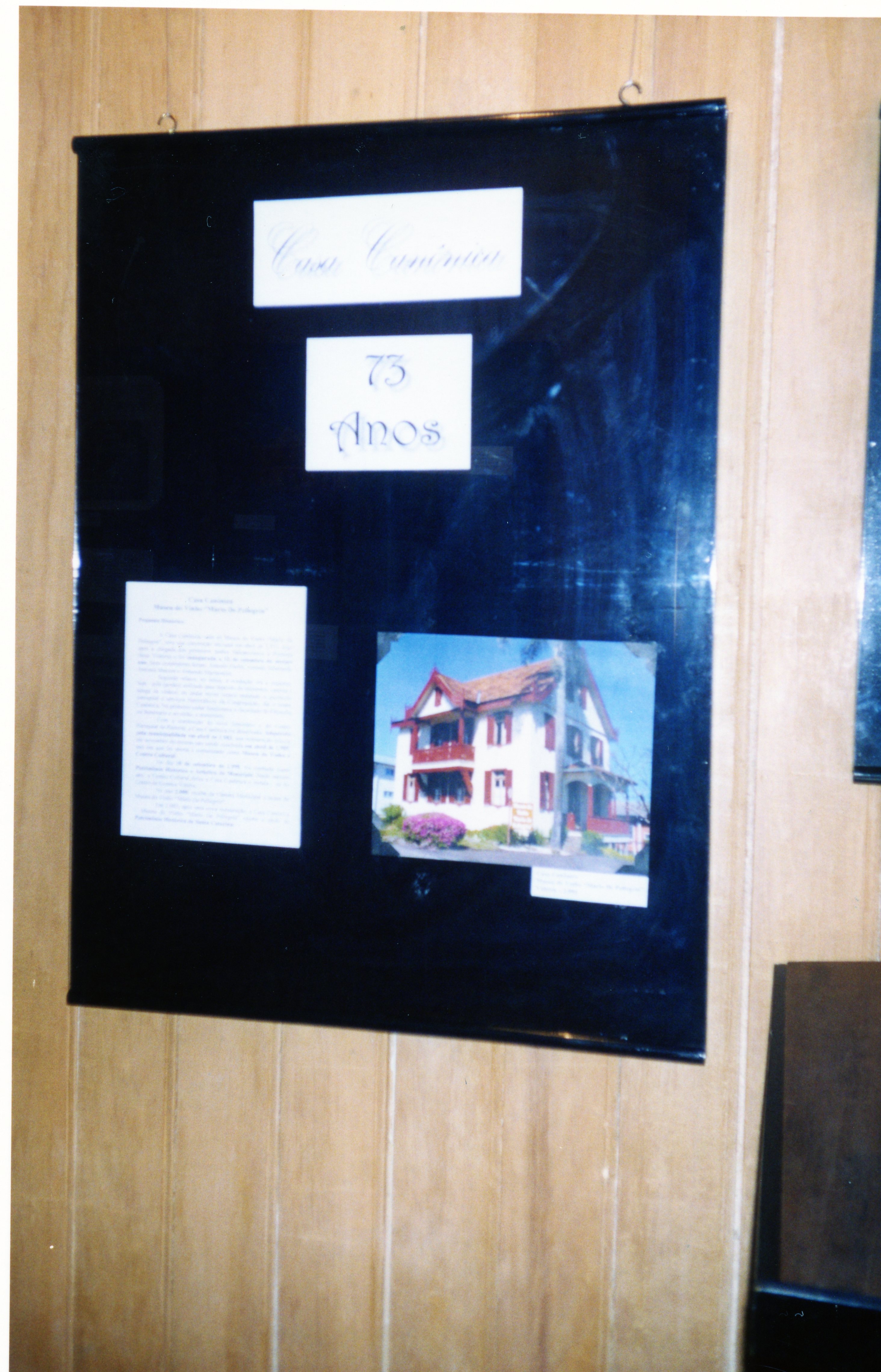 Um cartaz informativo fixado em uma parede de madeira, celebrando os 73 anos da Casa Canônica. O painel contém um texto descritivo e uma fotografia colorida de uma residência com arquitetura de estilo europeu, possivelmente em uma cidade de colonização imigrante como Videira.