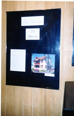 Um cartaz informativo fixado em uma parede de madeira, celebrando os 73 anos da Casa Canônica. O painel contém um texto descritivo e uma fotografia colorida de uma residência com arquitetura de estilo europeu, possivelmente em uma cidade de colonização imigrante como Videira.